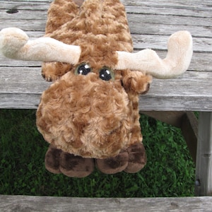 May include: A brown plush moose with large, white eyes and brown antlers. The moose is lying on a wooden surface.