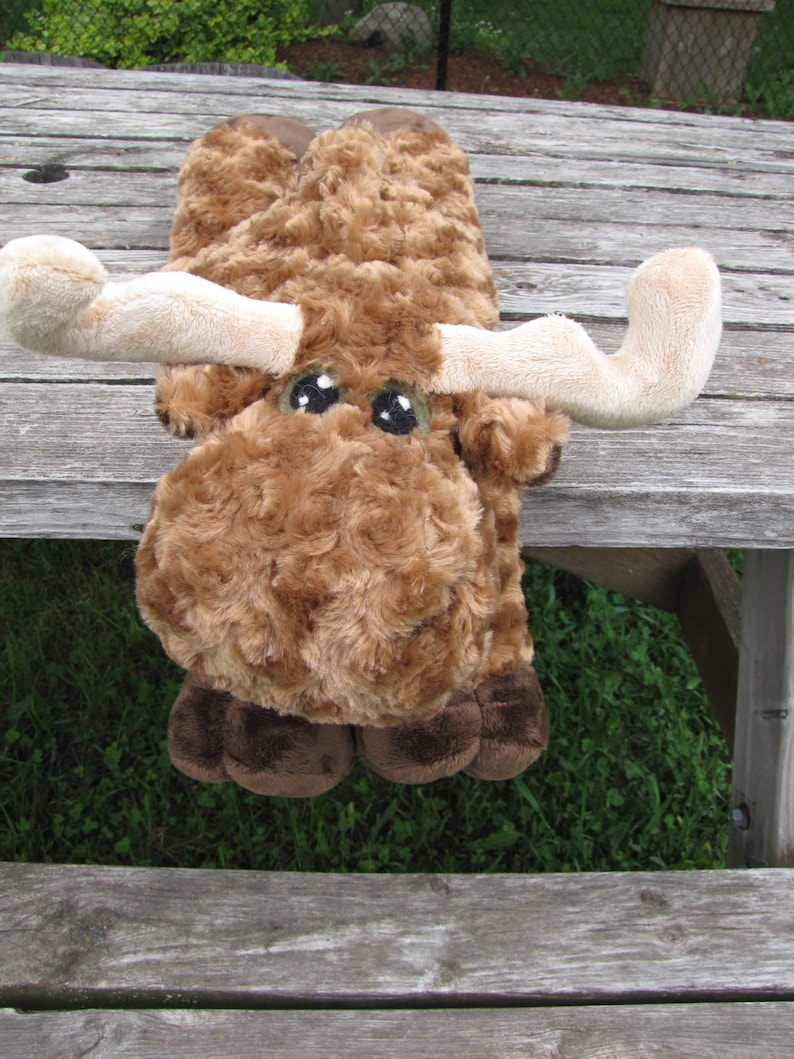 May include: A brown plush moose with large, white eyes and brown antlers. The moose is lying on a wooden surface.
