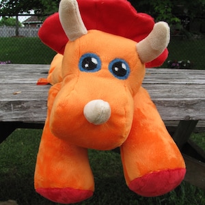 May include: Orange and red plush toy shaped like a triceratops dinosaur. The toy has large, blue eyes and a white horn on its nose.