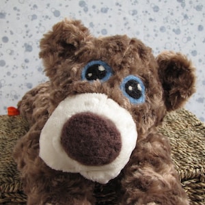 May include: A brown plush teddy bear with blue eyes and a white nose. The bear is lying on a woven basket.