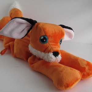 May include: Orange and white plush fox toy with black ears and blue eyes. The fox is lying on its belly with its tail curled up behind it.