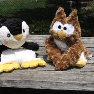 May include: Two plush stuffed animals: a penguin and an owl. The penguin is black and white with yellow feet and a yellow beak. The owl is brown with white and black eyes and a yellow beak. Both are sitting on a wooden surface.