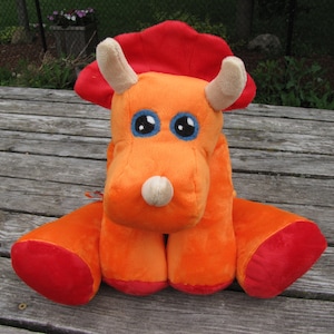 May include: Orange and red plush toy shaped like a triceratops dinosaur with large blue eyes.