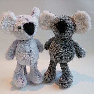 May include: Two plush koala toys, one gray and one light gray, with black noses and green eyes. The toys are standing on a white surface.