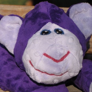 May include: Purple plush monkey toy with a white face and pink smile. The monkey has large, floppy ears and a soft, cuddly texture.