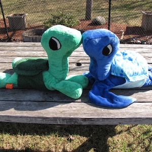 May include: Two plush turtle toys, one green and one blue, are lying on a wooden table. The green turtle has a brown shell and black eyes. The blue turtle has a light blue shell with a white pattern and black eyes.