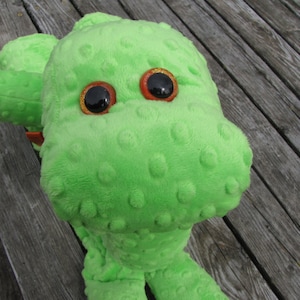 May include: A plush green dinosaur toy with large, glittery orange and black eyes. The soft toy has a textured surface with small, raised bumps. The dinosaur is lying on a weathered wooden surface.