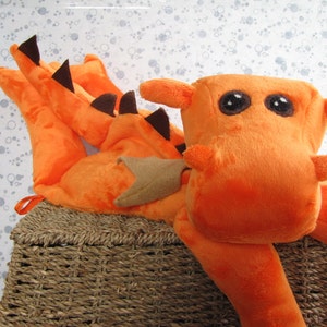 May include: Orange plush dragon toy with black eyes and a brown nose. The dragon is lying on a woven basket.