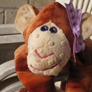 May include: A plush monkey toy with brown fur, tan face, and blue eyes. The monkey has a pink stitched smile and a purple ribbon with a floral pattern tied to its head. The toy is sitting on a white chair.