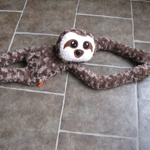 May include: A brown and white plush sloth toy with large, black eyes. The sloth is lying on its back with its arms outstretched.