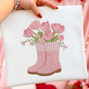 May include: White square pillow with an embroidered design of pink tulips in pink rain boots. The boots have a brown sole and a patterned cuff. The flowers and boots are in a detailed, stitched style.