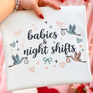 Può includere: Cuscino quadrato bianco con ricamo "babies & night shifts". Il design include cicogne, cuori, fiocchi e stelle in pastello blu, rosa e turchese. Il cuscino è tenuto da una persona.