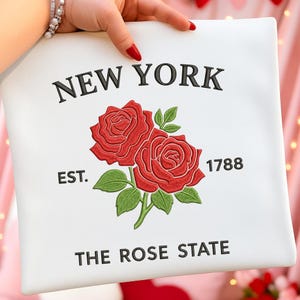 May include: White pillow with embroidered design featuring two red roses, green leaves, and the text "NEW YORK" above "THE ROSE STATE". The year "1788" is also included.