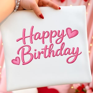 May include: A white rectangular pillow with the embroidered words "Happy Birthday" in pink script. Two pink hearts accent the text. The pillow is held by a hand with red nail polish.