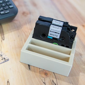 May include: Two white label tape cartridges for a label maker, in a beige plastic holder. The label tape is 12mm wide and 8m long, and is black on white. The label tape is labeled "Markomaier Laminated WHITE".