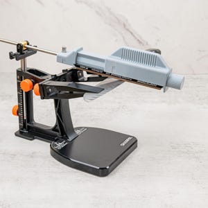 May include: A black metal knife sharpening system with orange adjustment knobs and a vertical scale. It features a light gray stone holder with a diamond sharpening stone attached, a horizontal guide rod, and a base labeled with the brand name SHARPAL.