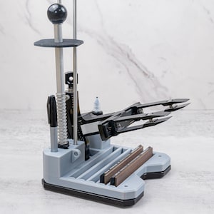 May include: A gray and black knife sharpening system with a black handle and a spring. The system has two brown sharpening stones and a black clamp to hold the knife. The base is gray and black.