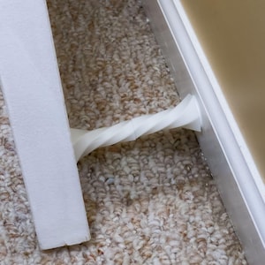 May include: A white, twisted plastic object, possibly a furniture component, is positioned between a white vertical support and a white baseboard. The object is set against a backdrop of beige carpet and a beige wall.