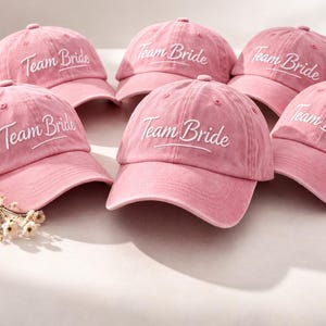 Peut inclure: Six casquettes de baseball roses avec le texte brodé "Team Bride" en écriture blanche. Les casquettes sont disposées sur une surface claire, avec un petit arrangement floral. Les casquettes sont de couleur rose unie.