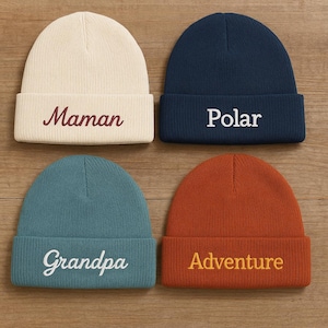 以下が含まれることがあります： クリーム、ネイビー、ティール、ラストオレンジの4色のニット帽。各帽子には折り返しがあり、刺繍されたテキストが施されています。「Maman」、「Polar」、「Grandpa」、「Adventure」。木製の表面に配置された帽子。