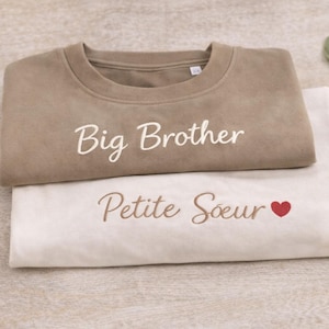 Può includere: Due felpe piegate, una color talpa e una bianco sporco. La felpa color talpa ha la scritta "Big Brother" ricamata in bianco. La felpa bianco sporco ha la scritta "Petite Soeur" ricamata in marrone con un cuore rosso.