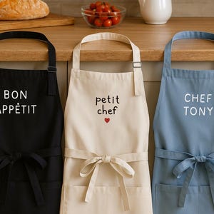 Peut inclure: Trois tabliers de cuisine, noir, crème et bleu. Le tablier noir porte l'inscription "BON APPÉTIT", le tablier crème "petit chef" avec un cœur, et le tablier bleu "CHEF TONY". Chaque tablier a une poche avant et une attache.