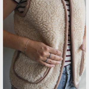 May include: A sleeveless beige sherpa waistcoat featuring a textured fleece exterior, a front zip fastening, and smooth brown trim along the edges. The waistcoat includes two front patch pockets and is worn over a white shirt with thin maroon stripes.