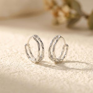May include: A pair of silver hoop earrings. Each earring has a textured, organic design with a double-hoop structure. The earrings are shown on a neutral, textured surface, with soft lighting.