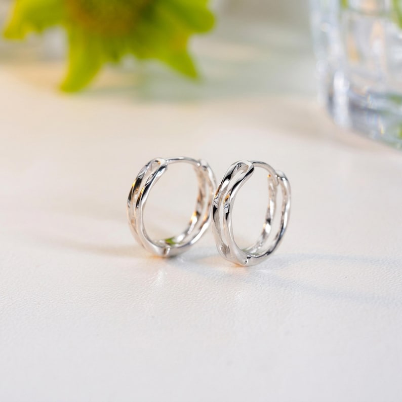 May include: A pair of silver hoop earrings. Each earring features a textured, twisted design. The earrings are presented on a white surface, with a blurred green and clear background.