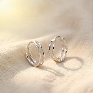 May include: A pair of silver hoop earrings with a distinctive layered design. The earrings are presented on a beige fabric, with soft lighting creating highlights and shadows. The hoops have a smooth, polished finish.