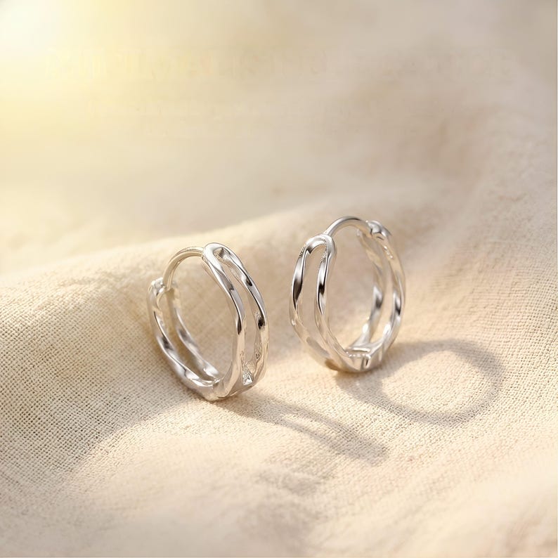 May include: A pair of silver hoop earrings with a distinctive layered design. The earrings are presented on a beige fabric, with soft lighting creating highlights and shadows. The hoops have a smooth, polished finish.
