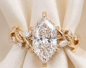 May include: A gold ring with a large, clear, marquise-cut diamond as the centerpiece. The band features a nature-inspired design with delicate leaves and smaller accent diamonds. The ring is set against a soft, white background.