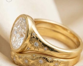May include: A gold ring set featuring a large oval-cut clear stone surrounded by a beaded border. The band and the matching contoured band are engraved with multiple starburst patterns, each accented with a small, embedded clear stone.