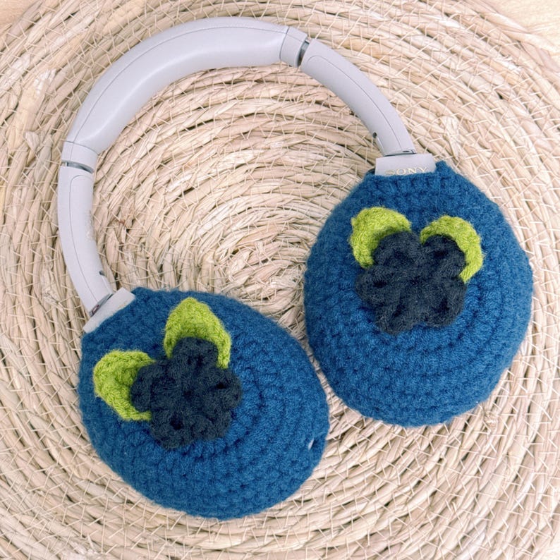 May include: Blue crochet headphone covers with a flower design in green and dark blue. The headphones have a gray headband and are resting on a woven, beige surface. The Sony brand is visible on the headphones.