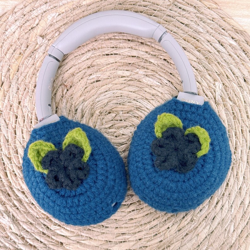 May include: Blue crochet headphone covers with a floral design in green and dark blue. The headphones have a gray headband and are resting on a woven mat. The ear covers are round and fully cover the headphone speakers.