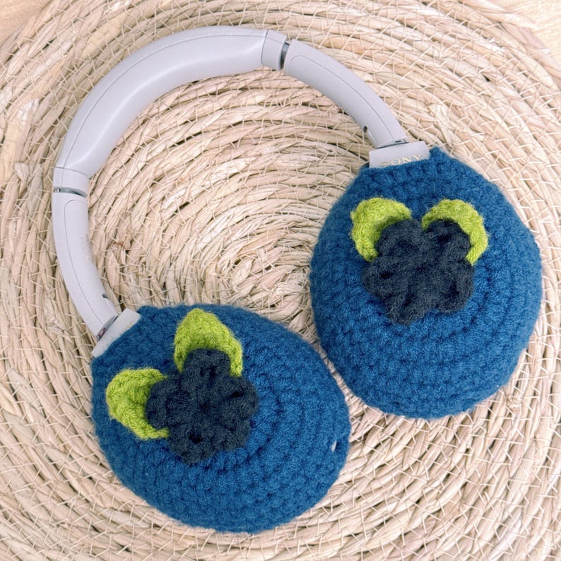 May include: A pair of gray headphones with blue crochet ear covers. Each cover features a dark blue and green flower design. The headphones rest on a woven, light brown surface.