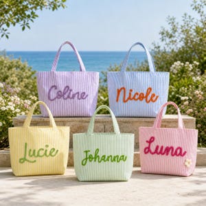 Peut inclure: Cinq sacs fourre-tout en seersucker aux couleurs pastel, chacun portant un prénom brodé en écriture cursive. Les sacs sont lavande avec 'Coline', bleu clair avec 'Nicole', jaune avec 'Lucie', vert clair avec 'Johanna' et rose avec 'Luna'.