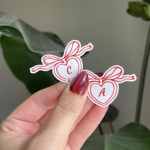 May include: Two heart-shaped stickers with red bows and outlines. One sticker has the letter "C" inside the heart, and the other has the letter "A". The stickers are white with red accents and are held by a hand.