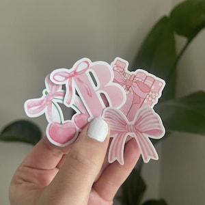 May include: A collection of pink stickers featuring a letter "B", cherries, and bows. The stickers have a pastel color scheme with a cute and feminine aesthetic. The stickers are die-cut and appear to be made of vinyl.