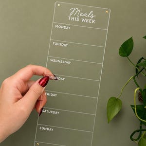 May include: A clear acrylic weekly meal planner board featuring white printed text that reads Meals THIS WEEK followed by the days of the week from Monday to Sunday. The rectangular board includes thin horizontal lines and small gold-toned hardware.