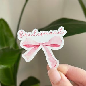 May include: A white sticker featuring a pink watercolour bow and the word "Bridesmaid" in cursive. The bow has a light pink centre and darker pink edges. The sticker is held by a person's hand.
