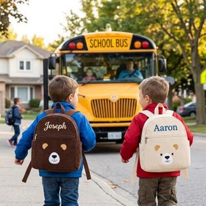 May include: Two children walk towards a yellow school bus, each wearing a backpack. One backpack is brown with "Joseph" embroidered on it, and the other is beige with "Aaron" embroidered. Both backpacks have a bear face design.