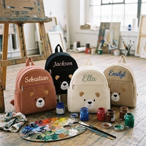 Puede incluir: Cuatro mochilas personalizadas con temática de oso en varios colores: marrón, negro, crema y beige. Cada mochila presenta un diseño de cara de oso y un nombre bordado en la parte delantera. Las mochilas se exhiben con materiales de pintura, incluyendo una paleta, pinceles y botes de pintura.