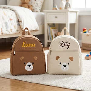 May include: Two corduroy backpacks, one brown and one cream, each with a bear face design. The brown backpack has "Luna" embroidered in yellow, and the cream backpack has "Lily" embroidered in brown. Both have a top handle and a front zipper.