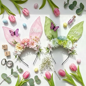 Puede incluir: Dos diademas de orejas de conejo de Pascua, una rosa y otra verde, decoradas con flores y mariposas. Las orejas rosas tienen el nombre "Mia" y las orejas verdes el nombre "Ivy". Huevos de Pascua y tulipanes completan la escena.