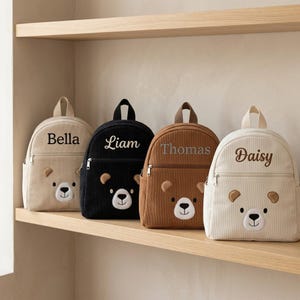 May include: Four corduroy backpacks in beige, black, brown, and cream, each with a bear face design and a name embroidered on the front: Bella, Liam, Thomas, and Daisy. The backpacks have a top handle and a front zip pocket.