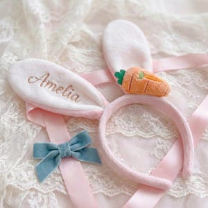 May include: A pink bunny ear headband with the name "Amelia" embroidered on one ear. A small orange carrot and a blue bow are also included. The headband is surrounded by pink ribbon and rests on a lace surface.
