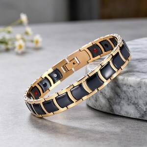 May include: A link bracelet featuring alternating gold-toned metal segments and smooth, glossy black rectangular inserts. The interior of the bracelet displays small circular accents in red, silver, and black colours, secured with a metal clasp.
