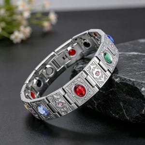 May include: A silver-toned link bracelet featuring rectangular segments with engraved floral patterns. The bracelet is accented with various oval-shaped coloured gemstones, including red, green, and blue, along with small embedded pink crystals.