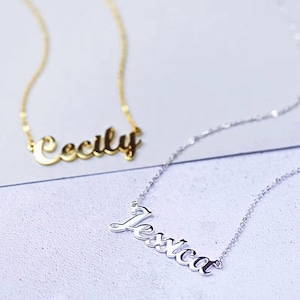 Könnte beinhalten: Zwei zarte Namensketten. Eine ist goldfarben und schreibt "Cecily" in einer Schreibschrift. Die andere ist silberfarben und schreibt "Jessica" in einem ähnlichen Stil. Beide Halsketten werden auf einer hellen Oberfläche präsentiert.
